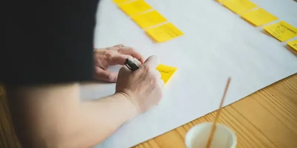 Person schreibt auf gelben Postit, im Hintergrund weitere Postits